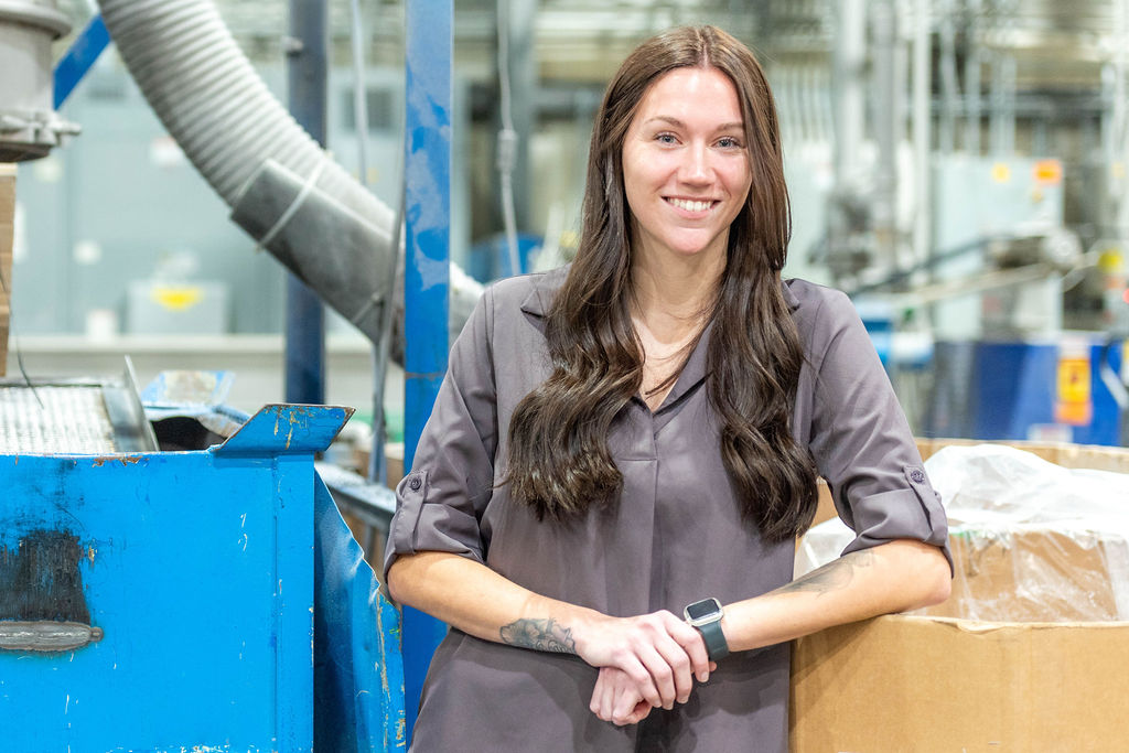 A woman with long brown hair, wearing a gray blouse and smartwatch, stands smiling in an industrial factory setting, leaning on a large cardboard box beside blue machinery and pipes.