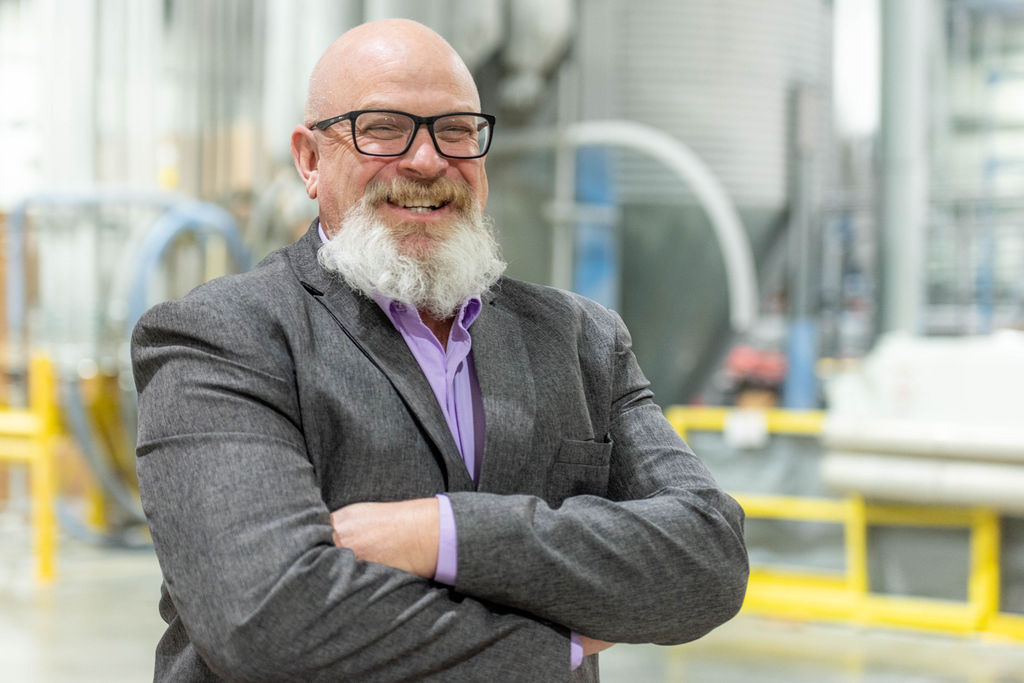 A smiling, bald man with a gray beard and glasses stands with arms crossed in an industrial or factory setting, wearing a gray suit jacket and a light purple shirt.