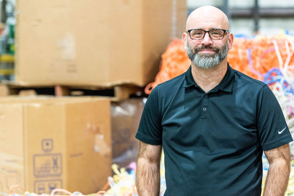 A smiling man with a bald head, beard, and glasses stands in front of large cardboard boxes and colorful tangled cords in what appears to be a warehouse or factory setting.