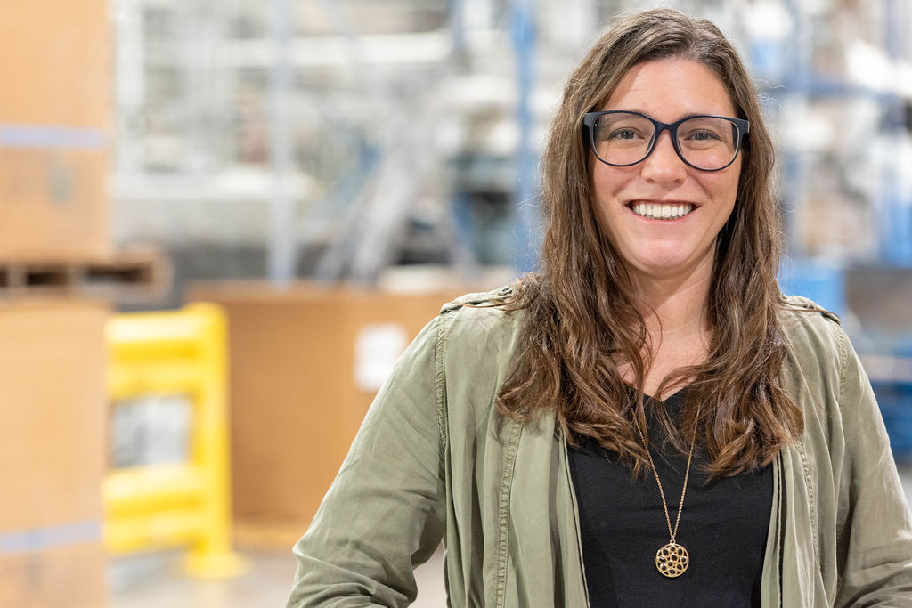 A woman with long brown hair and glasses smiles while standing in a warehouse. She wears a green jacket, black shirt, and a pendant necklace. The background is blurred with industrial shelves and yellow barriers.