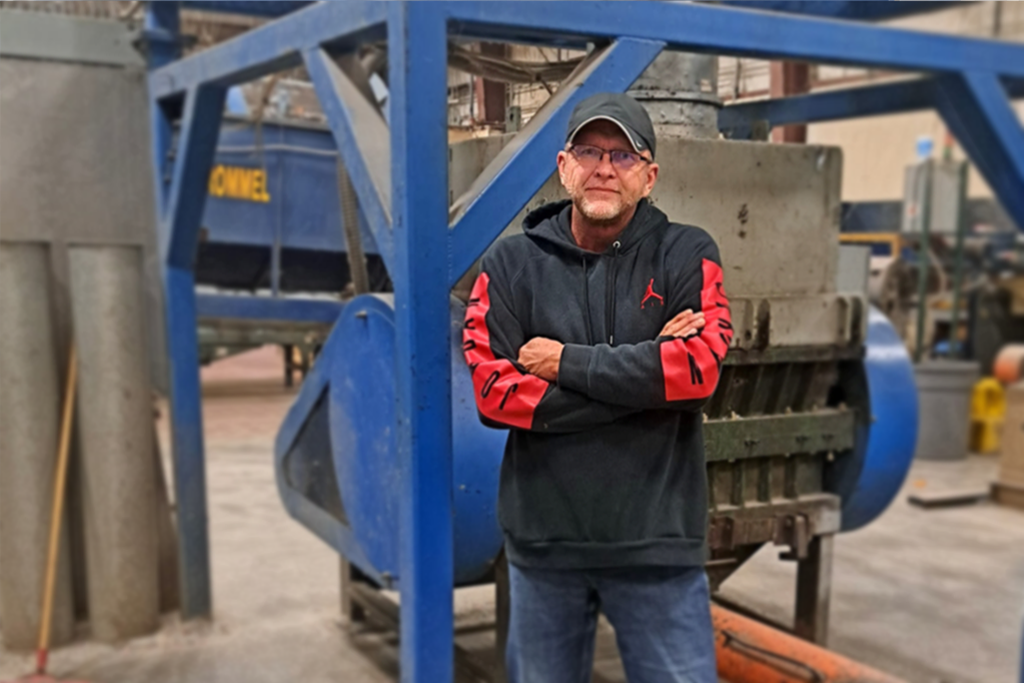 A man wearing glasses, a black cap, and a black hoodie with red sleeves stands with arms crossed in an industrial setting, surrounded by blue metal structures and heavy machinery.